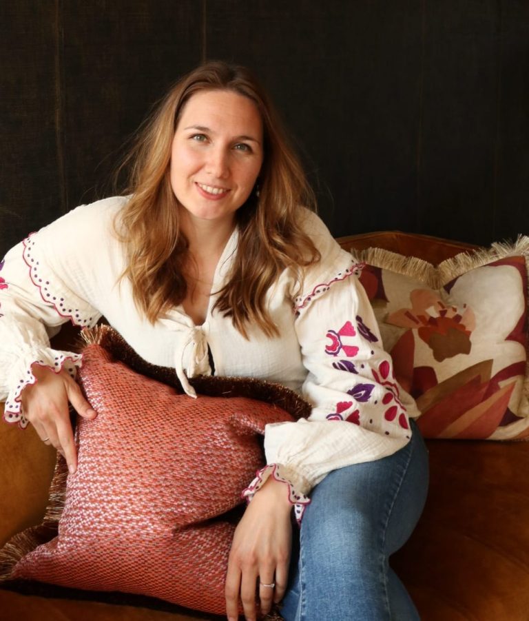 Vrouw met lang haar in een witte blouse, zittend op een bank met kussens.